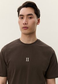 Ung man med kort mörkt hår som bär en mörkbrun t-shirt med en vit romersk siffra II på bröstet, neutral ljus bakgrund.