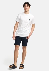 T-shirt bianca in cotone con un piccolo logo blu navy, abbinata a pantaloni corti blu navy e sandali neri con zeppa in sughero e design a due cinghie.
