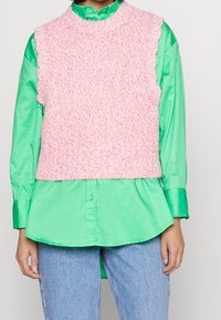 Henkilö, jolla on päällään kirkkaanvihreä nappipaita, pinkki neulottu hihaton liivi ja vaaleansiniset farkut.