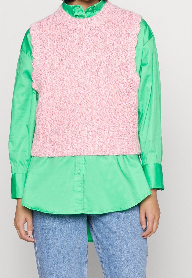 Henkilö, jolla on päällään kirkkaanvihreä nappipaita, pinkki neulottu hihaton liivi ja vaaleansiniset farkut.