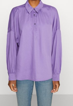 Person iført en løstsiddende, langærmet lavendelfarvet bluse med knappet krave og lyseblå jeans, stående mod en ensfarvet baggrund.