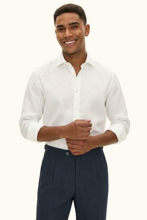 Smiling man wearing a white long-sleeve button-up shirt and dark blue pants, with hands clasped in front.