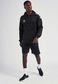 Zwarte hoodie met ritssluiting en een gestructureerde afwerking, met een logo op de borst en witte accenten op de schouders, gecombineerd met zwarte sportshorts.