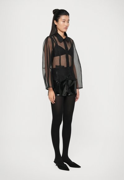 Čierna priesvitná košeľa na gombíky s dlhými rukávmi, prekrytá čiernym krátkym topom, v kombinácii s lesklými čiernymi šortkami a čiernymi pančuškami.