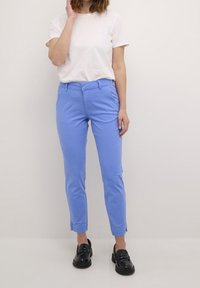 Pantalon ajusté bleu clair avec une texture lisse, jambes effilées, poches avant et fentes latérales à l'ourlet, associé à un t-shirt blanc et des chaussures noires.