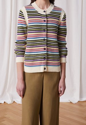 Personne portant un cardigan tricoté rayé multicolore avec des boutons noirs et un pantalon large beige, debout devant un fond de rideau blanc.