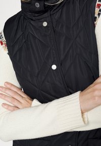 Schwarze gesteppte Weste mit hohem Kragen, Druckknopfverschluss und Rautenmuster, über einem cremefarbenen, gerippten Pulloverärmel getragen.