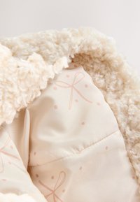 Krém színű műszőrme puha, bolyhos textúrával, amely egy sima belső bélést tár fel, melyet rózsaszín masnik és pöttyök mintája díszít.