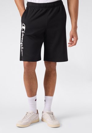 Hombre que lleva pantalones cortos deportivos negros con el logo blanco de Champion en la pierna izquierda, calcetines blancos y zapatillas color crudo, de pie sobre un fondo liso.