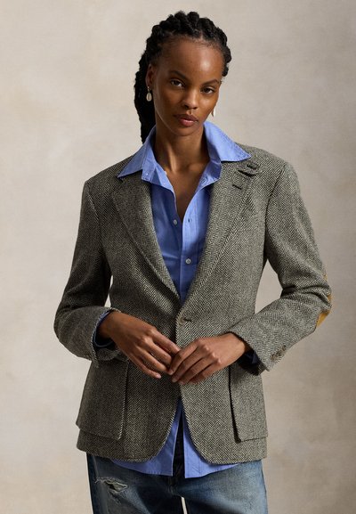 Femme aux cheveux tressés portant un blazer en chevrons sur une chemise bleue et un jean déchiré, debout avec les mains croisées à la taille.
