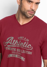 T-shirt bordeaux a V in tessuto morbido con stampa argentata che presenta le scritte "Athletic" e "Established 1986". Maniche corte, vestibilità comoda.