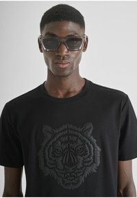 Crna majica s teksturiranim, uzdignutim uzorkom crnog tigra na prednjem dijelu. Model nosi prozirne naočale za sunce. Jednostavan, moderni dizajn.
