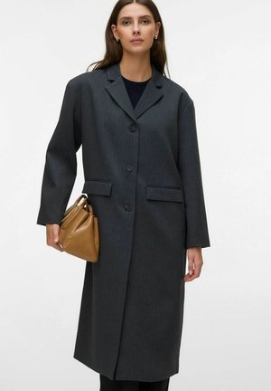 Femme portant un long manteau gris foncé boutonné tenant un sac à main marron sur un fond clair uni.