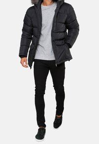 Svart puffjacka med avtagbar huva i päls, dragkedja och två framfickor. Bärs över en grå sweatshirt och svarta byxor.