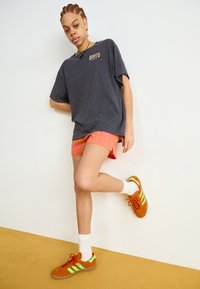 T-shirt oversized gris foncé avec logo, short à carreaux roses, chaussettes blanches, et baskets orange avec accents verts.