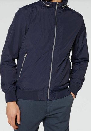 Mand iført en navyblå windbreakerjakke med lynlåslommer og hænderne i lommerne på mørkeblå bukser, stående foran en grå baggrund.