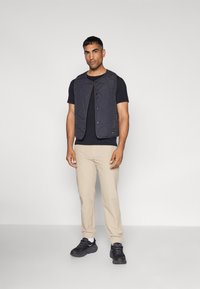 Gequilted donker vest over een zwart T-shirt, beige broek, en zwarte sneakers met witte accenten. Gladde texturen en drukknoopsluitingen zichtbaar.
