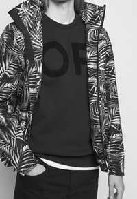 Giacca con cappuccio a stampa floreale con un motivo di foglie di palma in bianco e nero, indossata sopra un maglione scuro con un design testuale morbido "OF".