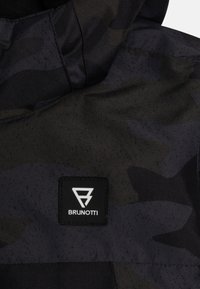 Svart och mörkgrå kamouflagejacka med en slät textur. Har en märkt svart fyrkantig lapp med vitt logotyp. Sömnadsdetaljer syns.