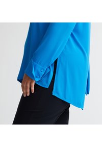 Blaue Bluse mit langen Ärmeln und glatter Textur, mit geknöpften Manschetten und seitlichen Schlitzen, über einer schwarzen Hose getragen.