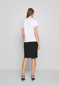 Vit t-shirt med kort ärm och rund halsringning, prydd med en liten logotyp på baksidan. Bärs tillsammans med en figurnära svart kjol och sandaletter med remmar.