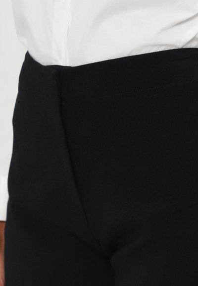 Zwarte broek met een gladde textuur en een getailleerde pasvorm. De tailleband ligt plat, in contrast met een wit shirt dat gedeeltelijk zichtbaar boven de broek is.