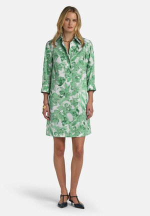 Frau trägt ein knieumspielendes grünes und weißes Blumenkleid mit dreiviertel Ärmeln, steht mit schwarzen T-Riemchen-Absätzen vor weißem Hintergrund.