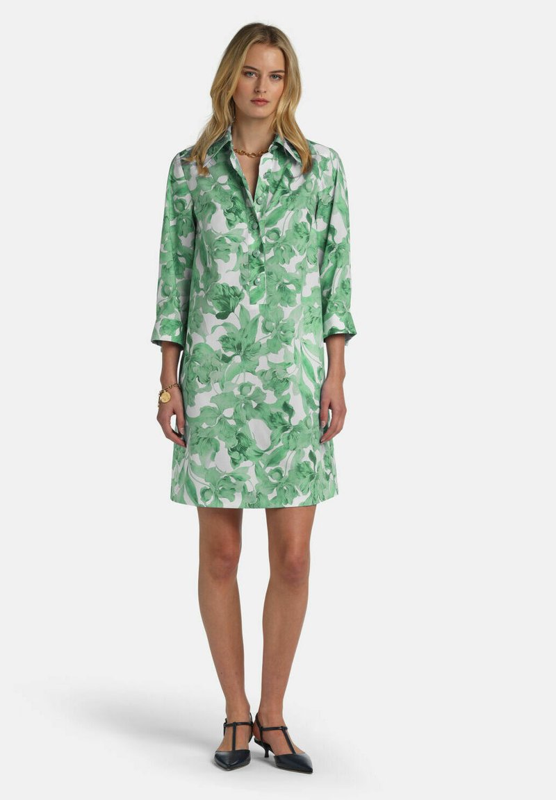 Frau trägt ein knieumspielendes grünes und weißes Blumenkleid mit dreiviertel Ärmeln, steht mit schwarzen T-Riemchen-Absätzen vor weißem Hintergrund.