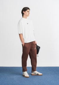 Camiseta blanca de gran tamaño con un pequeño logo negro, pantalones deportivos marrones con bolsillos laterales y zapatillas blancas con detalles en negro y marrón.