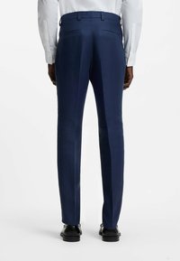 Homme portant un pantalon habillé bleu marine ajusté et des chaussures habillées noires, debout les mains le long du corps, dos à un fond blanc.
