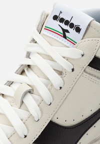 Baskets en cuir crème et noir, avec une languette blanche portant une étiquette de logo. Les surfaces texturées et les lacets plats blancs complètent le design.