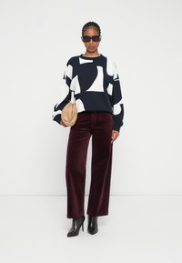 Jersey azul marino con formas abstractas blancas, combinado con pantalones de pana burdeos y botas de tacón negro afiladas. Sosteniendo un bolso de mano beige.