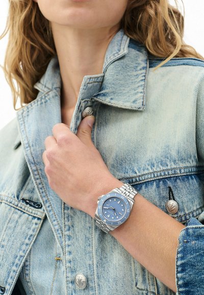 Isik helerohelises teksajakis reguleerib vasaku käega kraet, kandes hõbedast käekella sinise sihverplaadi ja metallist rihmaga.