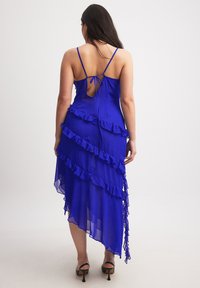 Asymmetrical blue chiffon dress with ruffled detailing, adjustable spaghetti straps, and an open back design. Black heels complete the look.