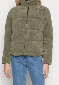 Giacca puffer in corduroy verde oliva con zip frontale, indossata con jeans in denim blu, mostrata dalla zona del collo ai fianchi.