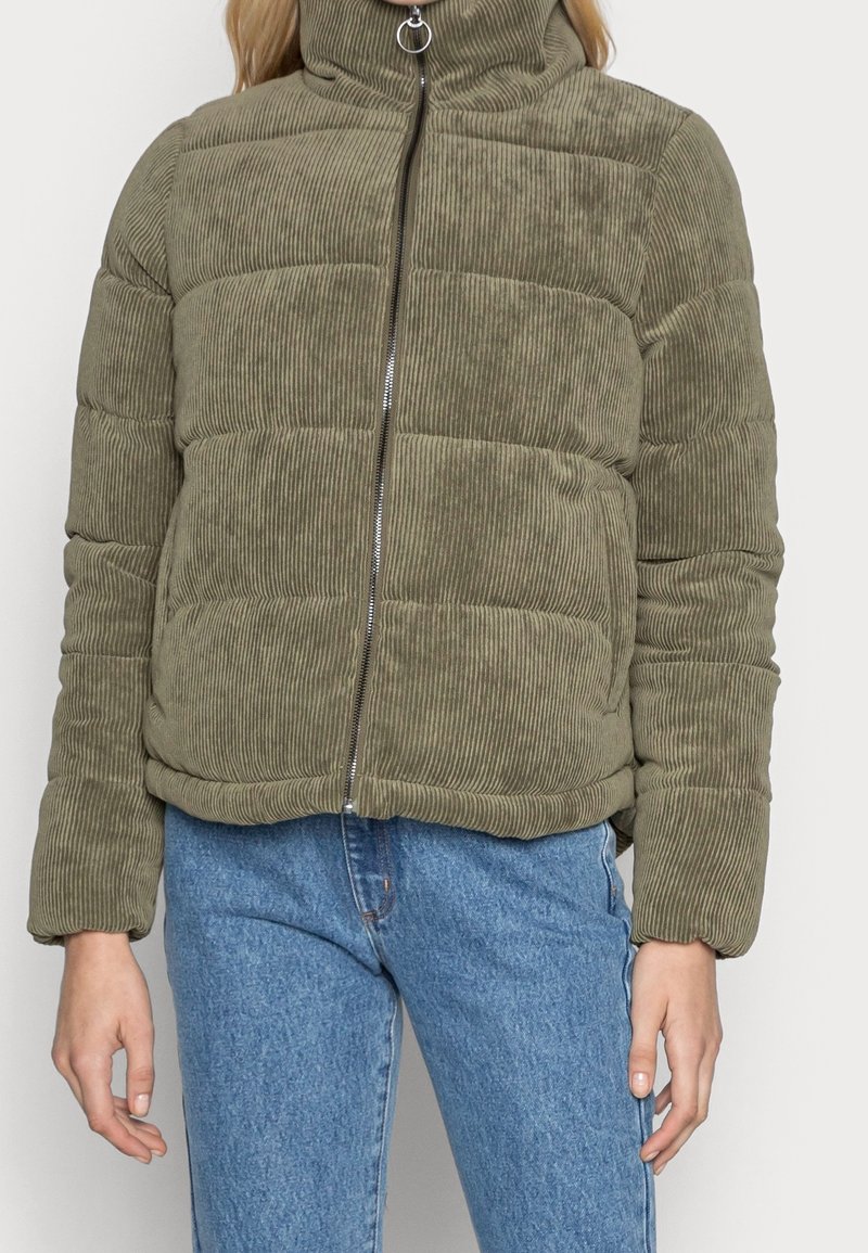 Giacca puffer in corduroy verde oliva con zip frontale, indossata con jeans in denim blu, mostrata dalla zona del collo ai fianchi.