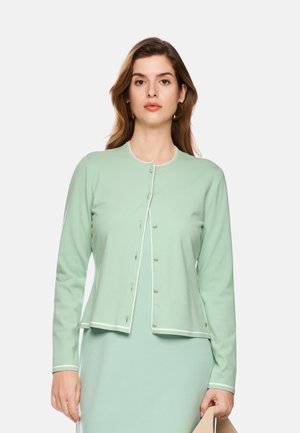 Woman with light brown hair wearing a light green buttoned cardigan over a matching dress holding a beige hat against a plain white background.