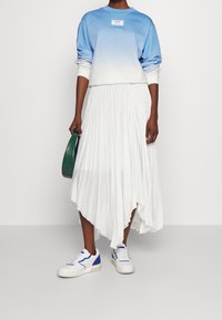 Sudadera azul degradada con un logo, falda asimétrica blanca plisada y zapatillas blancas con detalles azules; bolso verde en mano.