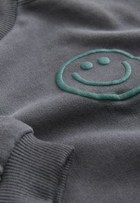 Tessuto a maglia grigio con polsini a costine e un design di faccina sorridente sollevata di colore teal sulla superficie.