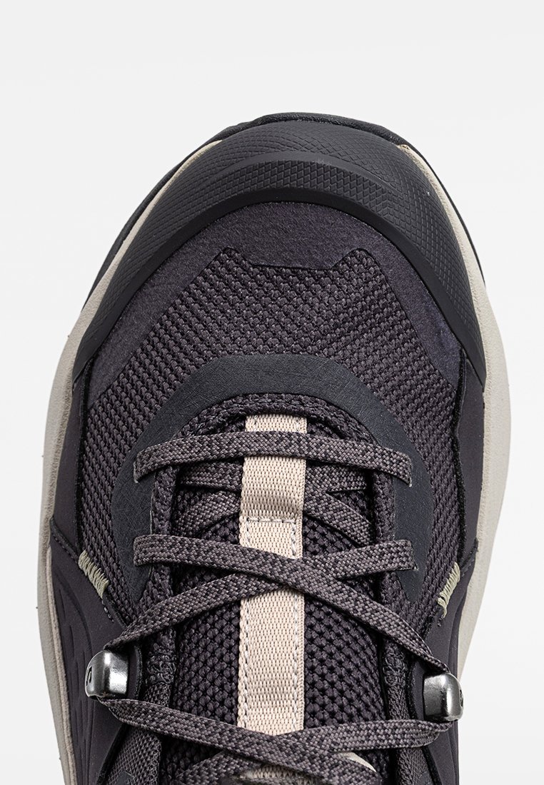 Nahaufnahme eines schwarzen und grauen Sportschuhs mit Mesh-Gewebe, beiger Mittelstreife, überkreuzten Schnürsenkeln und strukturiertem Gummifußschutz.