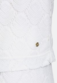 Biała tkanina o teksturze z wzorem liści, z małym złotym emblematem. Gładkie wykończenie z warstwowym detalem u dołu.