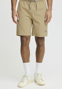Beige swim shorts with an elastic drawstring waist, side pockets, and a small logo detail on the lower leg. Worn with white socks and sneakers.