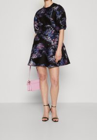 Svart klänning med blommönster i lila och blått, korta puffärmar, utsvängd kjol. Åtföljd av en liten rosa handväska och svarta klacksandaler.