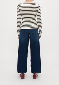 Gestreiftes Langarmoberteil in Creme und Marineblau, kombiniert mit weit geschnittenen dunkelblauen Jeans. Rote Schuhe und Socken sind am Knöchel sichtbar.
