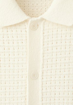 Gros plan sur un cardigan tricoté blanc cassé avec un motif gaufré texturé, une patte de boutonnage à l'avant et un col plat.
