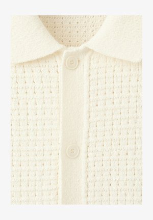 Gros plan sur un cardigan tricoté blanc cassé avec un motif gaufré texturé, une patte de boutonnage à l'avant et un col plat.