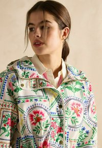 Pelerină de ploaie cu un model floral, având o bază albă, accente vibrante de roz, verde și albastru, dotată cu glugă și accesorii de culoare aurie.