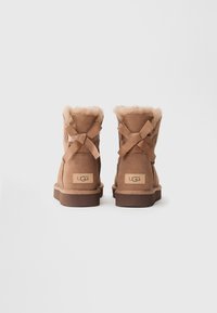 Bottines en daim marron avec un intérieur pelucheux, dotées d'un lien à ruban à l'arrière et d'une semelle en caoutchouc plate. Logo UGG sur le côté.