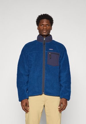 Jachetă din fleece albastru cu fermoar în față, guler înalt și un buzunar pe piept cu o pată de material bleumarin. Material moale, texturat.