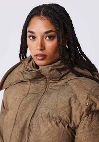 Chaqueta acolchada marrón con cuello alto, con textura arrugada y detalles sutiles de costura. Cierre con cremallera y botones a presión en el hombro.